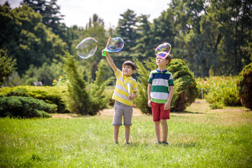 Fototapeta premium Boy blowing soap bubbles while an excited kid enjoys the bubbles. Happy teenage boy and his brother in a park enjoying making soap bubbles. Happy childhood friendship concept