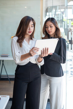 Image of two young Asian woman stand holding tablet by the window at the office.