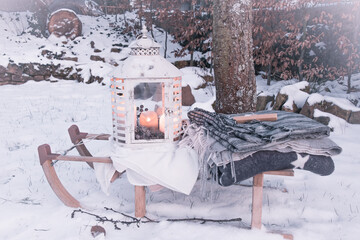 White vintage lantern with candles and various woolen blankets on a sled in a snowy garden