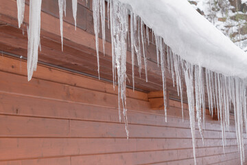 Icicles on the roof of the building. White snow build-up. Wooden facing of the front wall. Concept of danger of falling icicles and snow from the roof.