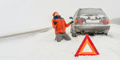 Angry young man in an orange jacket is kneeling on a snow-covered road. He is in a state of stress because his car is broken. Winter travel and recreation, road problems and assistance concepts.Banner
