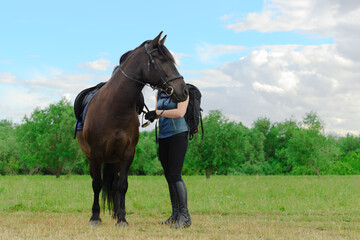 Caucasian horse rider is standing next to her mare and adjusting the stirrup of saddle.