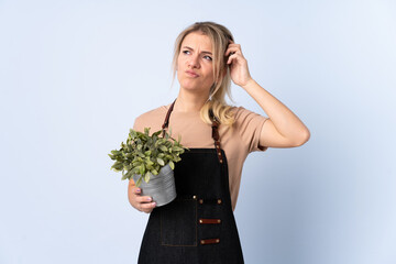 Blonde gardener woman holding a plant over isolated background having doubts and with confuse face expression