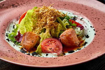 Appetizing warm salad with baked salmon, cherry tomatoes, lemongrass and white sauce, served in a white plate on a wooden background. Restaurant food