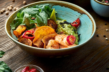 Appetizing mexican appetizer - avocado guacamole with shrimps, vegetable chips and cilantro in a bowl on a wooden background. Close up view