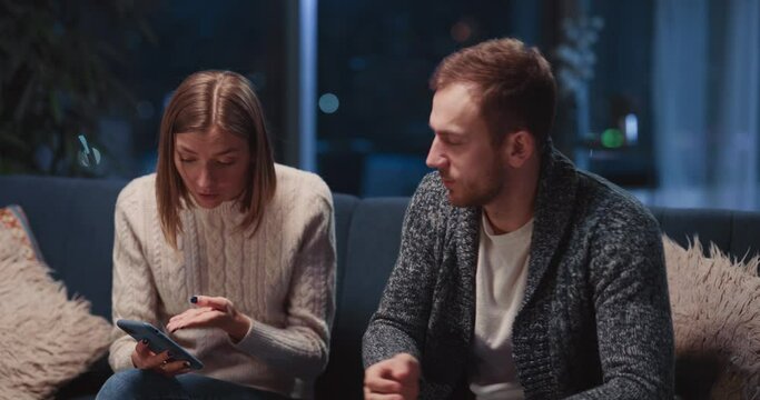 Caucasian young married couple making up conflict in their apartment. Angry young woman fell jealous about her husband reading messages on his smartphone.