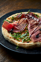 Wood-fired pizza with jamon, arugula, red sauce and balsamic with crispy sides, served on a black plate on a wooden background. pizzette a kind of Italian pizza