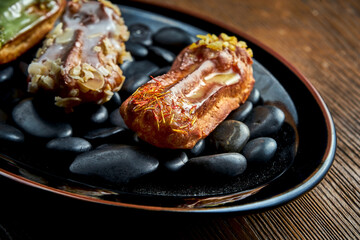 Set of assorted French custard eclairs with custard and assorted toppings served on a black plate on a wooden background. Restaurant food.