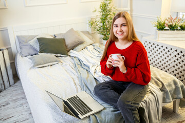 Beautiful young woman working on a laptop sitting on the bed at home, she is drinking coffee and smiling. Work from home during quarantine. Girl in a sweater and jeans at home on the bed