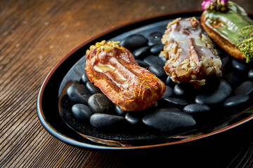 Set of assorted French custard eclairs with custard and assorted toppings served on a black plate on a wooden background. Restaurant food.