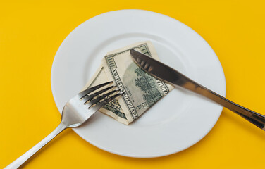 A hundred-dollar bill lies on a white ceramic plate near a fork and a knife on a yellow cotton tablecloth. The concept donation of business lunch, banking and financial life rentier.