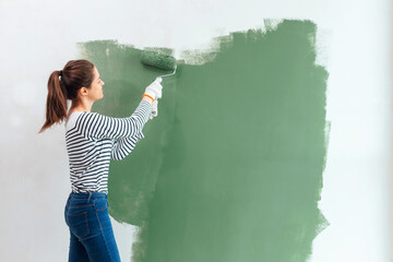 Young woman is painting the wall into green with paint roller in new house