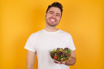 young handsome Caucasian man holding a salad bowl against yellow wall laughing.