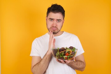 young handsome Caucasian man holding a salad bowl against yellow wall keeps palms pressed together in front of her having regretful look, asking for forgiveness. Forgive me please.