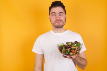 young handsome Caucasian man holding a salad bowl against yellow wall being nervous and scared biting lips looking camera with impatient expression, pensive.
