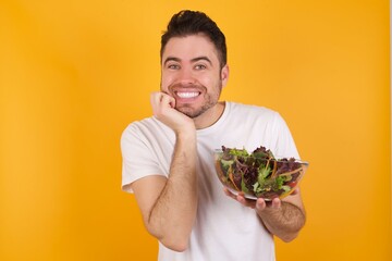 Dreamy young handsome Caucasian man holding a salad bowl against yellow wall keeps hands pressed together under chin, looks with happy expression, has toothy smile.