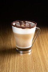 Latte art coffee with syrup, milk and chocolate in a transparent glass with a metal handle on a wooden table