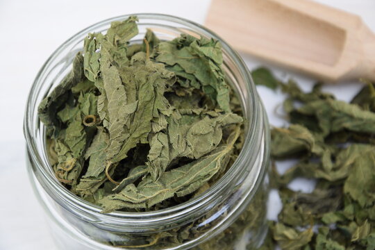 Dried nettle leaves in glass jar closeup on white wooden background, wooden shovel