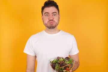 young handsome Caucasian man holding a salad bowl against yellow wall puffing cheeks with funny face. Mouth inflated with air, crazy expression.