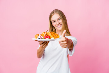 Young russian woman eating a waffle isolated with thumbs ups, cheers about something, support and respect concept.