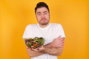 young handsome Caucasian man holding a salad bowl against yellow wall bitting his mouth and looking worried and scared crossing arms, worry and doubt.
