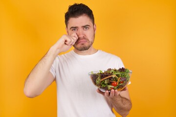 Unhappy young handsome Caucasian man holding a salad bowl against yellow wall crying while posing at camera whipping tears with hand.