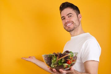 young handsome Caucasian man holding a salad bowl against yellow wall Inviting to enter smiling natural with open hands. Welcome sign.