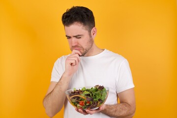 Sad young handsome Caucasian man holding a salad bowl against yellow wall feeling upset while spending time at home alone staring at camera with unhappy or regretful look.
