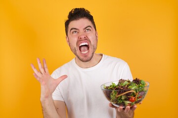 young handsome Caucasian man holding a salad bowl against yellow wall crying and screaming. Human emotions, facial expression concept. Screaming, hate, rage.