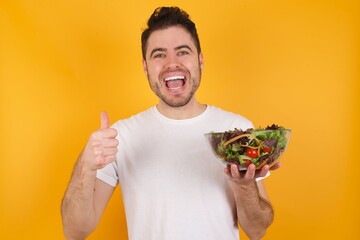 Optimistic young handsome Caucasian man holding a salad bowl against yellow wall showing thumbs up with positive emotions. Quality and recommendation concept.