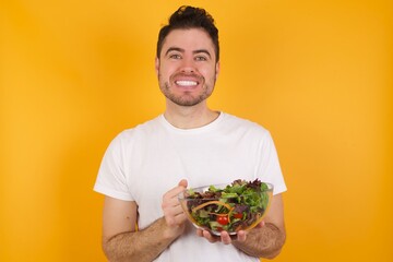 Happy young handsome Caucasian man holding a salad bowl against yellow wall stands against orange studio wall keeps hands on heart, swears be loyal, expresses gratitude. Honesty concept.