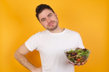 Funny frustrated young handsome Caucasian man holding a salad bowl against yellow wall holding hands on waist and silly looking at awkward situation.