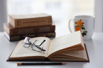 Old books, glasses, pencil and cup of coffee on white table background against window light with copy space