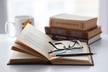 Fototapeta premium Old books, glasses, pencil and cup of coffee on white table background against window light with copy space. Vintage cozy warm mockup