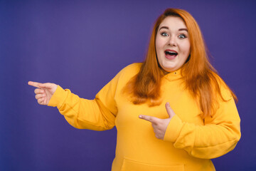 Excited fat woman with red hair pointing at copy space isolated over blue background