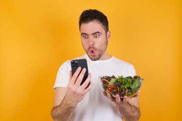 young handsome Caucasian man holding a salad bowl against yellow wall looks with bugged eyes, holds modern smart phone, receives unexpected message from friend, reads reminder.