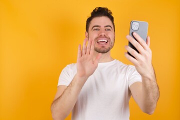 Portrait of happy friendly young handsome Caucasian man wearing white t-shirt against yellow wall taking selfie and waving hand, communicating on video call, online chatting.