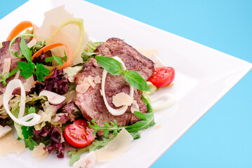 Warm salad with veal, tomatoes, cheese, lettuce, onion on a white plate on a blue background. close-up. photo for clipping. for the menu