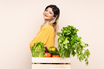 Teenager farmer girl with freshly picked vegetables in a box isolated on beige background laughing