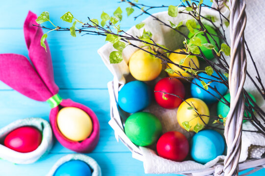 Easter eggs in the shape of rabbit ears, a basket with colorful Easter eggs and tree branches with green leaves, on a blue wooden background, selective focus, tinted image