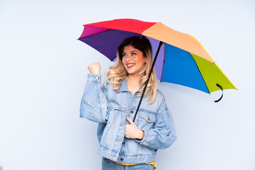 Teenager russian girl holding an umbrella isolated on blue background celebrating a victory