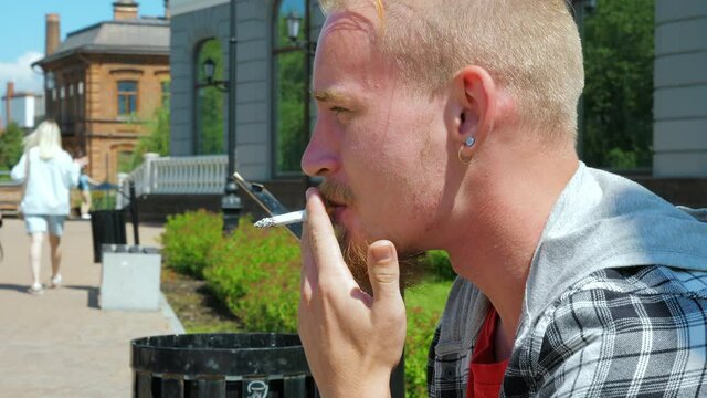 An anxious man and looking at women passing by. A young man is sitting on the bench and smoking nervously. A concept of stalking ex-partner, or perversion
