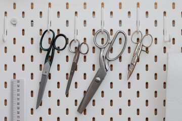 Tailor scissors in different sizes and forms hanging on hooks of perforated white wall