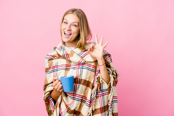 Young russian woman wrapped in a blanket drinking coffee winks an eye and holds an okay gesture with hand.