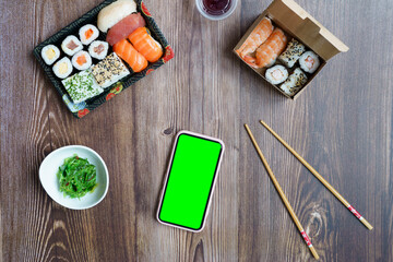 Mock up mobile with green screen on a wooden table with sushi food in take-away containers.