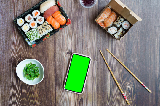 Mock up mobile with green screen on a wooden table with sushi food in take-away containers.