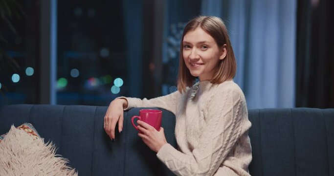 Porrait of cute beautiful young caucasian woman holdign cup with coffee resting inside apartment cozy home smiling satisfied to camera.