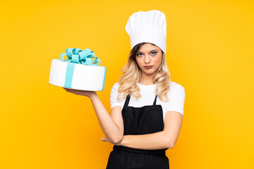 Teenager girl pastry holding a big cake isolated on yellow background keeping arms crossed