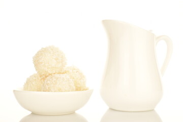 Several round sweets with coconut flakes in a ceramic white plate with a milk jug, close-up, isolated on white.