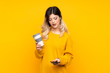 Teenager girl isolated on yellow background holding coffee to take away and a mobile